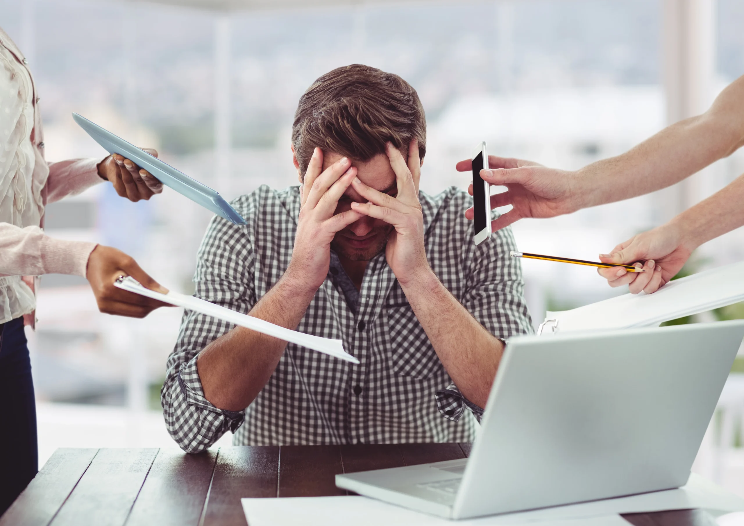 a young man sits at his computer with his hands over his face, clearly stressed, as several hands hold work-related items towards him.